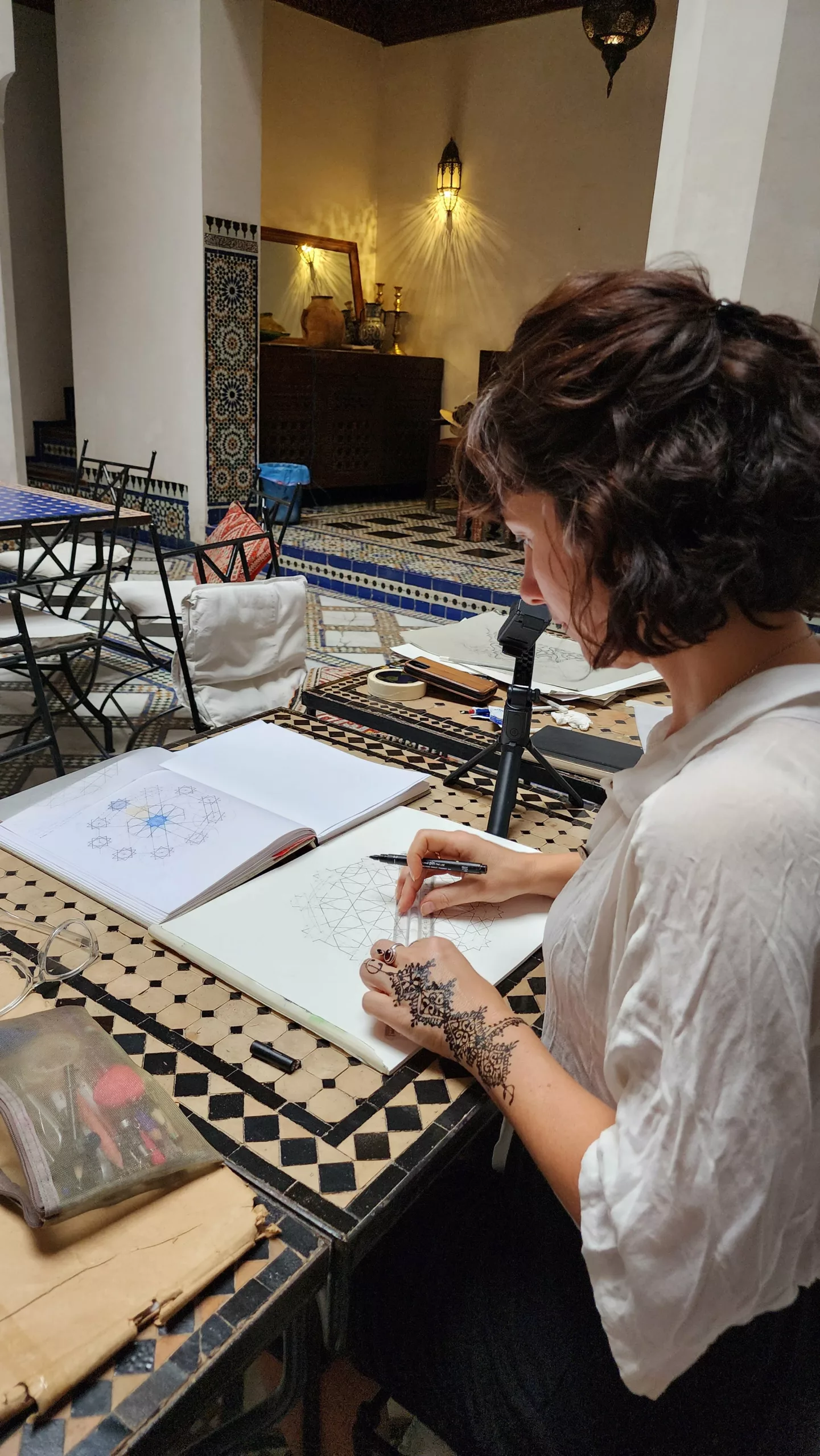 Mulher com henna na mão desenhando geometria sagrada à noite no pátio de um riad, com câmera em tripé registrando o caderno